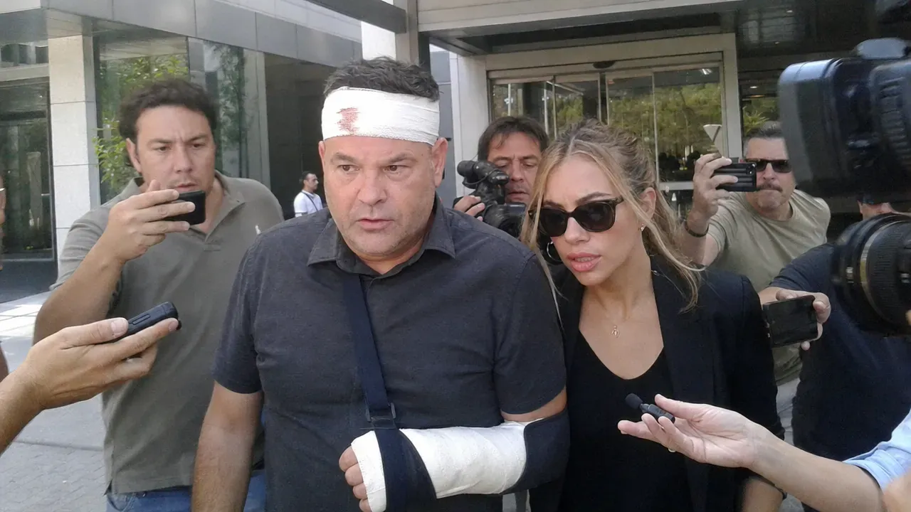 José Elías Navarro sale del hospital con la cabeza vendada y el brazo en cabestrillo, rodeado de periodistas con micrófonos y cámaras.